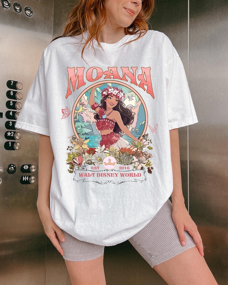 May include: White t-shirt featuring the word "MOANA" in pink lettering above a circular graphic. The graphic depicts a woman in a floral crown and traditional attire, surrounded by flowers and butterflies. The text "WALT DISNEY WORLD" is below the graphic.