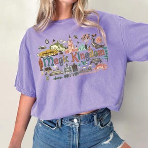May include: A lilac-colored t-shirt with a graphic that says "Magic Kingdom" in green and blue. The graphic includes illustrations of a castle, a boat, and other theme park elements. The shirt has short sleeves and a relaxed fit.