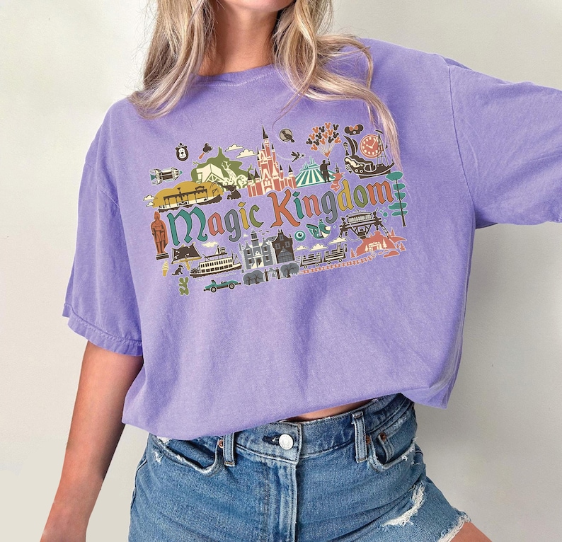 May include: A lilac-colored t-shirt with a graphic that says "Magic Kingdom" in green and blue. The graphic includes illustrations of a castle, a boat, and other theme park elements. The shirt has short sleeves and a relaxed fit.