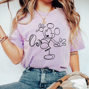 May include: A light purple t-shirt featuring a black line drawing of Mickey Mouse with the text "Oh Boy". The shirt is a crew neck style and has short sleeves. The brand name "ORCHID" is printed on the bottom left.