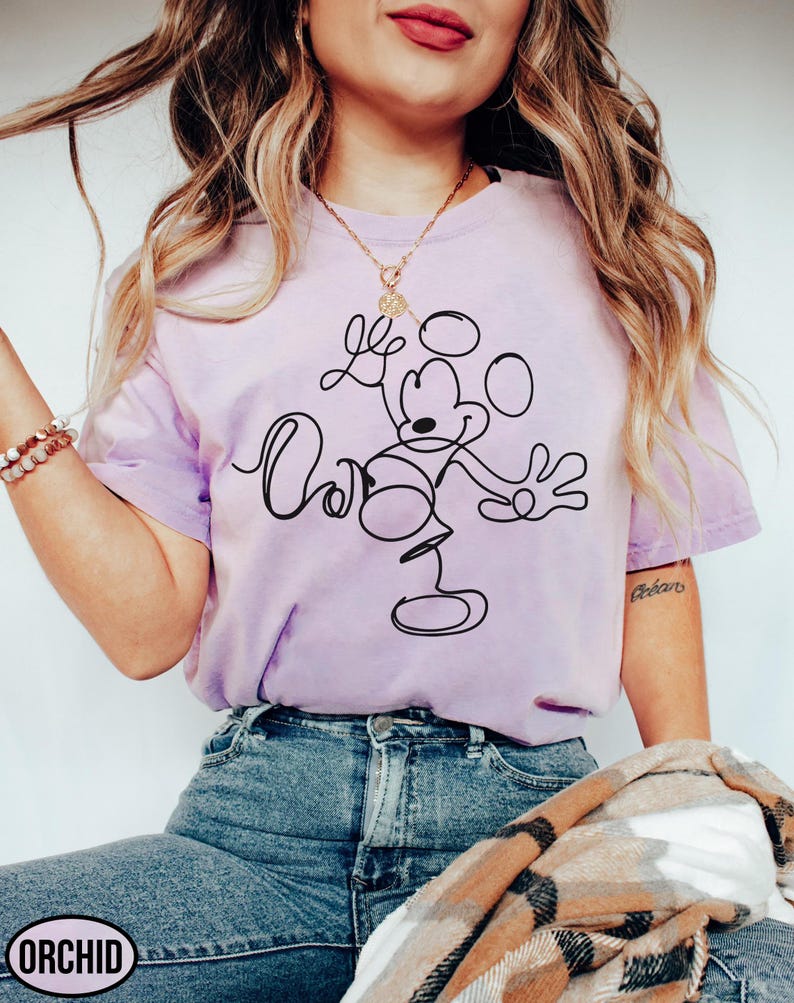 May include: A light purple t-shirt featuring a black line drawing of Mickey Mouse with the text "Oh Boy". The shirt is a crew neck style and has short sleeves. The brand name "ORCHID" is printed on the bottom left.