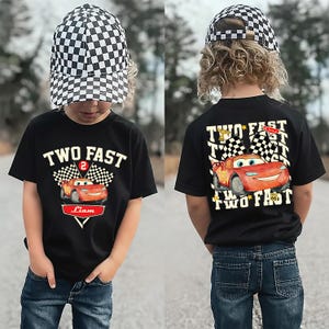 May include: Black t-shirt featuring a red race car graphic and the text "TWO FAST" and "Liam". The outfit includes a black and white chequered baseball cap. The back of the shirt also has the race car and the words "TWO FAST".