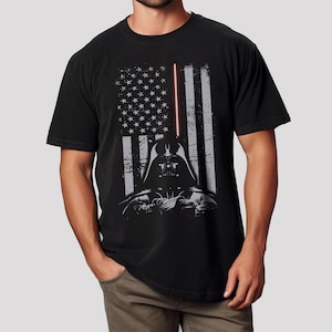 May include: Black t-shirt featuring a distressed American flag design with Darth Vader's helmet and a red lightsaber. The graphic is in shades of gray and white, creating a vintage look. The shirt is made of a soft material.