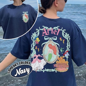May include: Navy blue t-shirt featuring a design from "The Little Mermaid." The back of the shirt displays the name "Ariel" in pink script above a framed image of Ariel, with other characters. The front has a smaller version of the design.