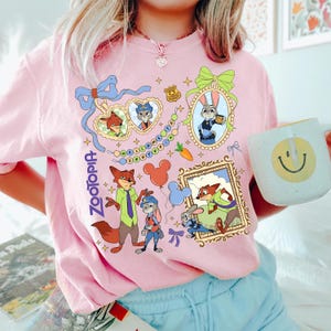 May include: Pink Zootopia-themed sweatshirt featuring cartoon characters Nick Wilde and Judy Hopps. The design includes framed portraits, Mickey Mouse balloons, and the text "Welcome to Zootopia". A white mug with a yellow smiley face is held in the image.