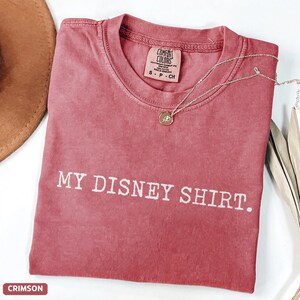 May include: Crimson-colored t-shirt with the text "MY DISNEY SHIRT." A silver necklace with a pendant rests on the shirt. The shirt has a crew neck and a tag that says "COMFORT COLORS."