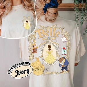 May include: Ivory-colored t-shirt featuring a design from Beauty and the Beast. The front has a small oval design with Belle in a yellow dress. The back has a larger design with Belle, the Beast, and other characters. The text "Belle" is at the top.
