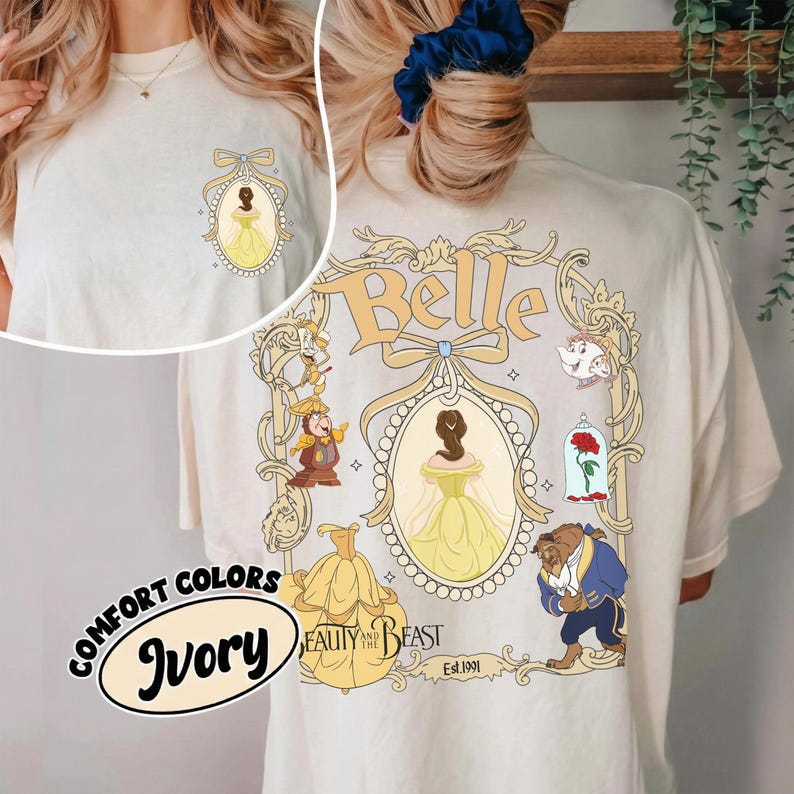May include: Ivory-colored t-shirt featuring a design from Beauty and the Beast. The front has a small oval design with Belle in a yellow dress. The back has a larger design with Belle, the Beast, and other characters. The text "Belle" is at the top.