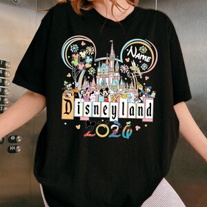 Op de afbeelding: Zwart T-shirt met een kleurrijke Disneyland-afbeelding. Het ontwerp bevat het Disneyland-logo, een kasteel en verschillende Disney-personages. Het jaar "2020" wordt ook weergegeven. Het shirt heeft een ronde hals en korte mouwen.