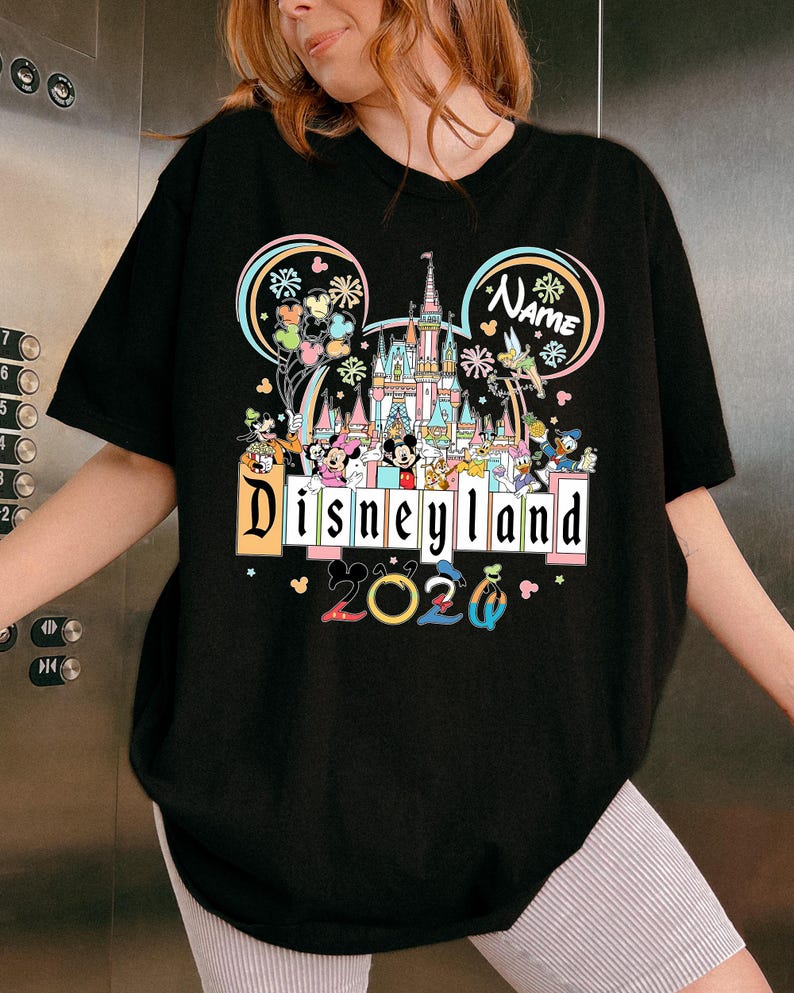 Op de afbeelding: Zwart T-shirt met een kleurrijke Disneyland-afbeelding. Het ontwerp bevat het Disneyland-logo, een kasteel en verschillende Disney-personages. Het jaar "2020" wordt ook weergegeven. Het shirt heeft een ronde hals en korte mouwen.