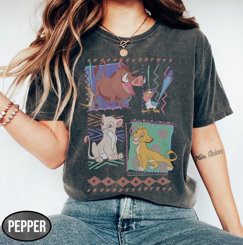 May include: Dark gray t-shirt featuring a colorful graphic of characters from The Lion King. The design includes Timon, Pumbaa, Simba, and Nala, set against patterned backgrounds. The shirt has a vintage, washed look.