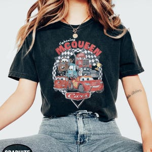 Puede incluir: Camiseta negra con un dise&ntilde;o gr&aacute;fico vintage de Rayo McQueen y otros personajes de la pel&iacute;cula Cars. El dise&ntilde;o incluye el texto "Lightning McQueen" y "Cars", sobre un fondo de bandera a cuadros.