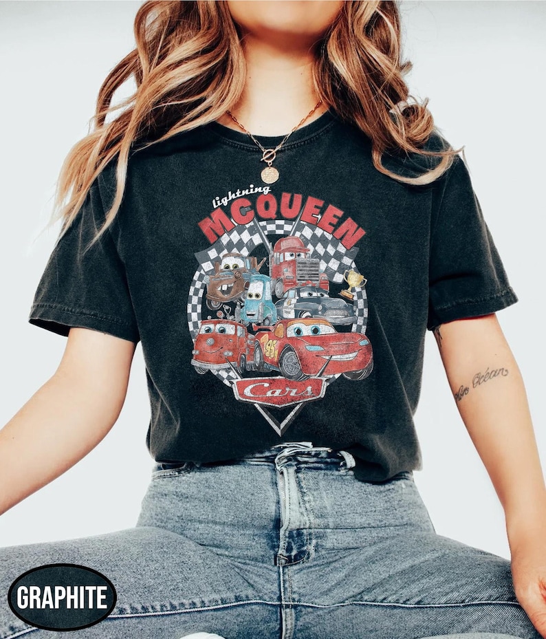 Puede incluir: Camiseta negra con un dise&ntilde;o gr&aacute;fico vintage de Rayo McQueen y otros personajes de la pel&iacute;cula Cars. El dise&ntilde;o incluye el texto "Lightning McQueen" y "Cars", sobre un fondo de bandera a cuadros.