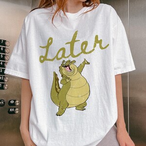 May include: White t-shirt featuring a cartoon alligator with the word "Later" in a green, cursive font. The alligator is light green and yellow, with its mouth open and arms raised. The shirt is a casual, oversized fit.