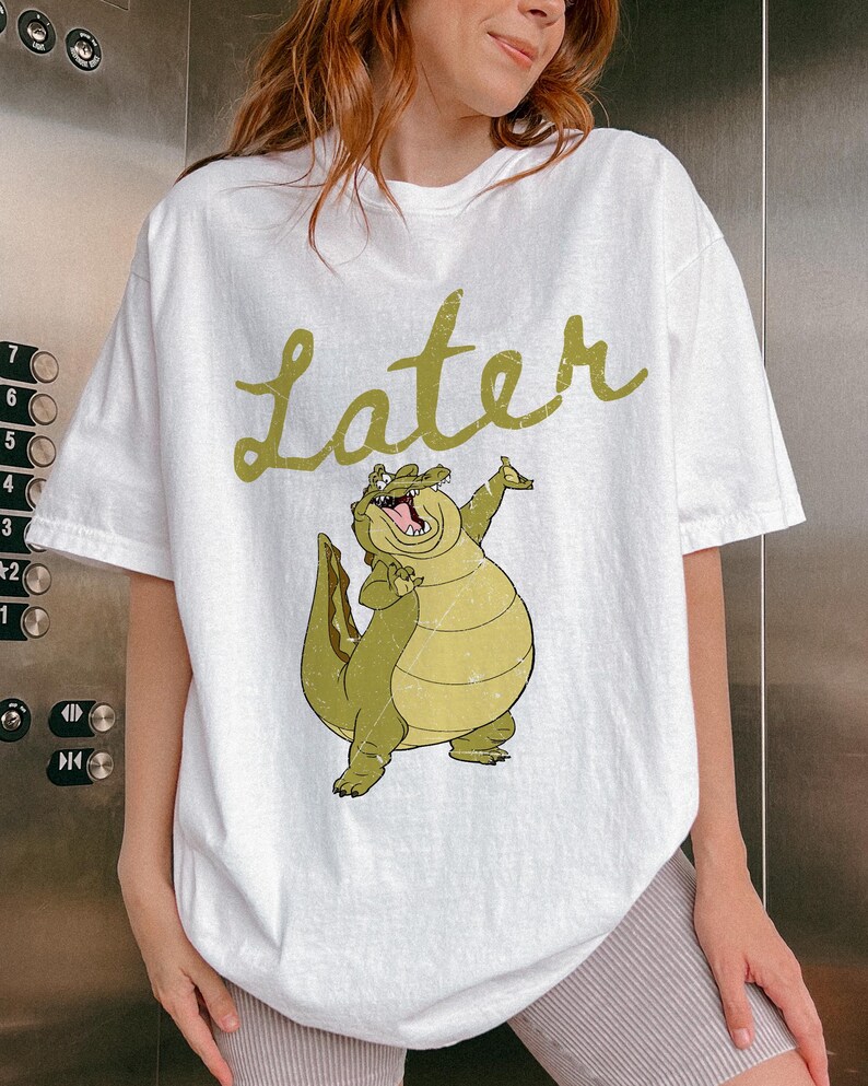 May include: White t-shirt featuring a cartoon alligator with the word "Later" in a green, cursive font. The alligator is light green and yellow, with its mouth open and arms raised. The shirt is a casual, oversized fit.
