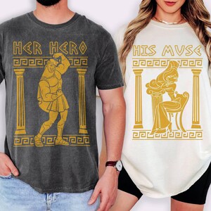 May include: Two t-shirts, one dark gray and one off-white, featuring Greek-inspired illustrations. The dark gray shirt says "HER HERO" with a gold image of a muscular figure. The off-white shirt says "HIS MUSE" with a gold image of a woman.