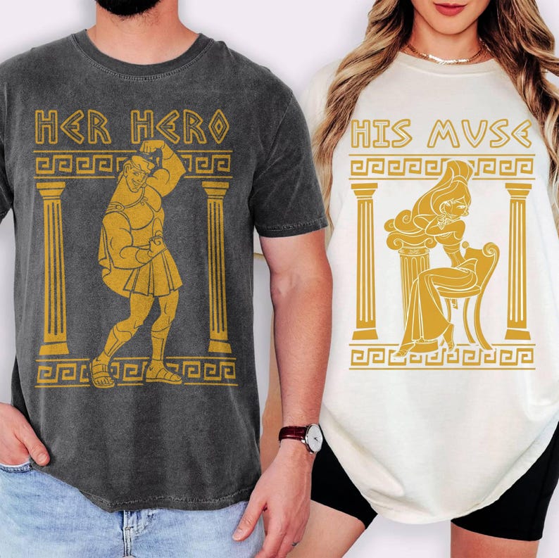 May include: Two t-shirts, one dark gray and one off-white, featuring Greek-inspired illustrations. The dark gray shirt says "HER HERO" with a gold image of a muscular figure. The off-white shirt says "HIS MUSE" with a gold image of a woman.