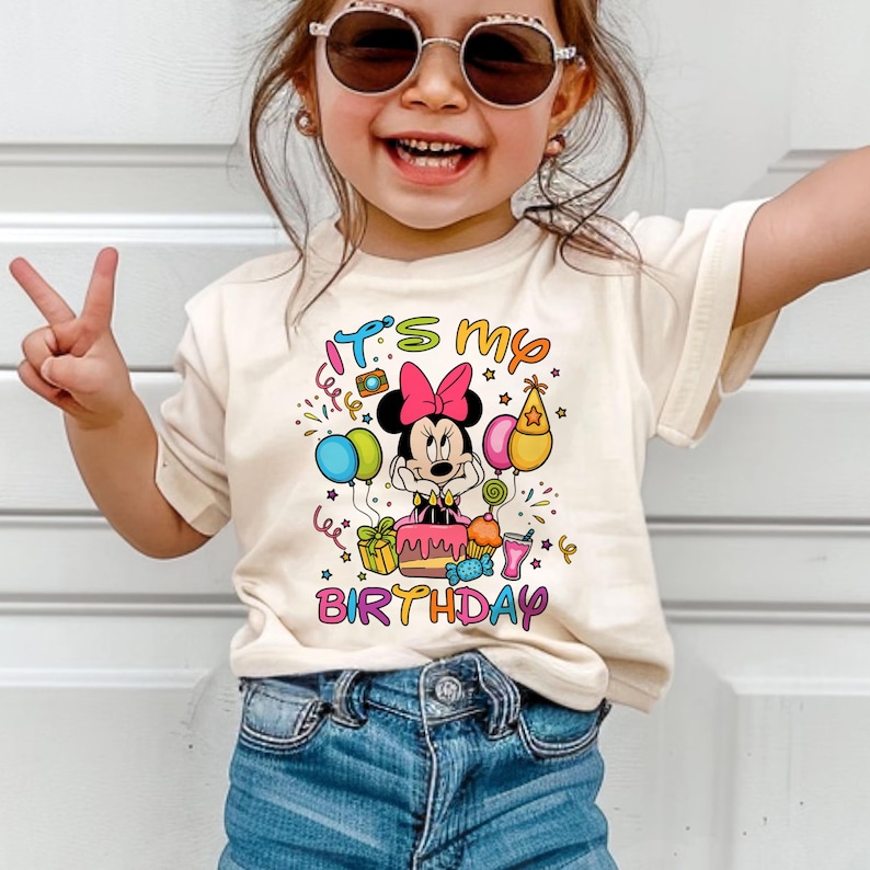 May include: A cream-colored t-shirt featuring a colorful graphic of Minnie Mouse with a pink bow, a birthday cake, balloons, and the text "It's my birthday". The shirt is paired with blue denim jeans.