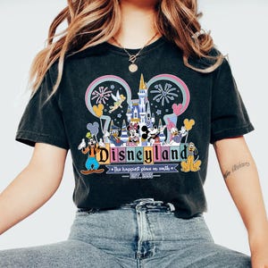 Może przedstawiać: Czarny t-shirt z kolorową grafiką Disneylandu. Projekt zawiera logo Disneylandu, kultowe postacie, takie jak Myszka Miki, i zamek. Widoczny jest r&oacute;wnież tekst "Disneyland" i "The happiest place on earth".