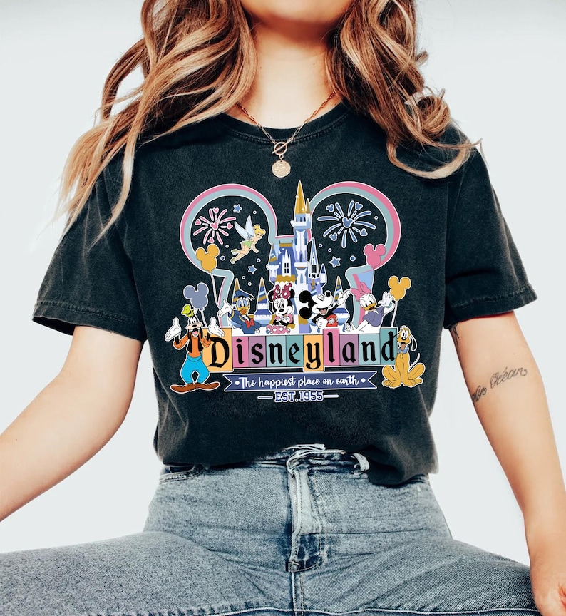 Może przedstawiać: Czarny t-shirt z kolorową grafiką Disneylandu. Projekt zawiera logo Disneylandu, kultowe postacie, takie jak Myszka Miki, i zamek. Widoczny jest r&oacute;wnież tekst "Disneyland" i "The happiest place on earth".