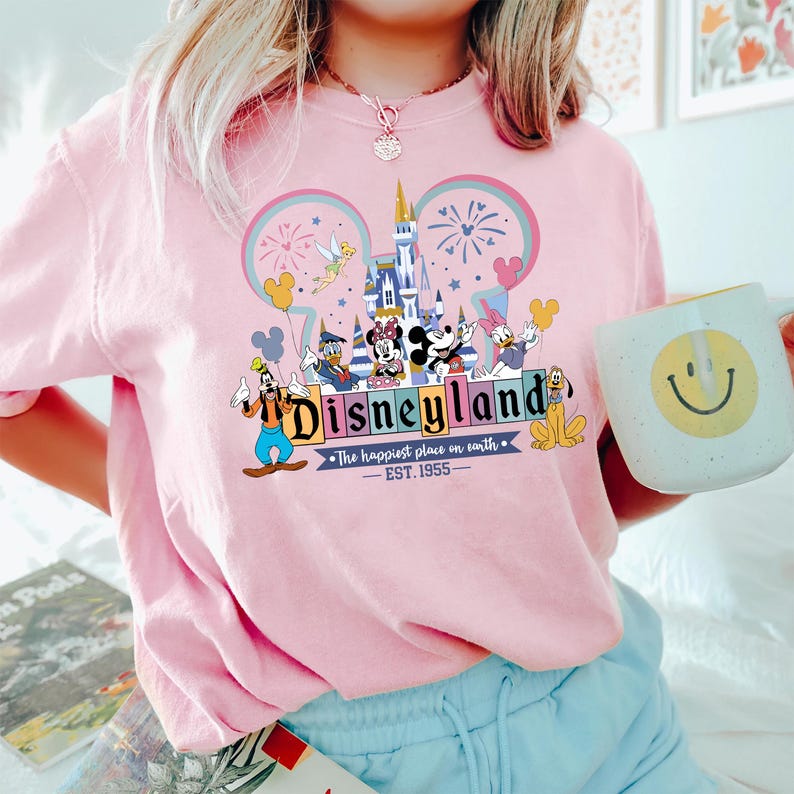 Może przedstawiać: R&oacute;żowa bluza z okrągłym dekoltem z grafiką Disneyland z Myszką Miki, Minnie, Donaldem i innymi postaciami Disneya. Grafika zawiera tekst "Disneyland" i "The happiest place on earth - EST. 1955". Widoczny jest r&oacute;wnież biały kubek z ż&oacute;łtą uśmiechniętą buzią.