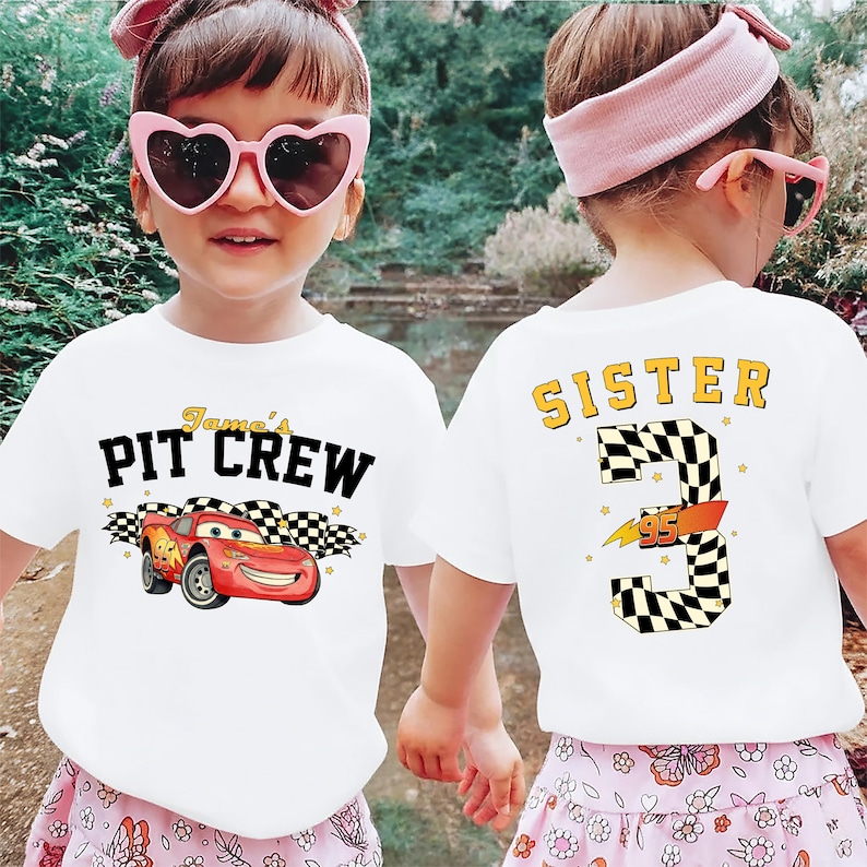 May include: White t-shirts with a race car theme. One shirt features "James's PIT CREW" with a red race car and checkered flags. The other shirt says "SISTER 3 95" with a checkered number 3.