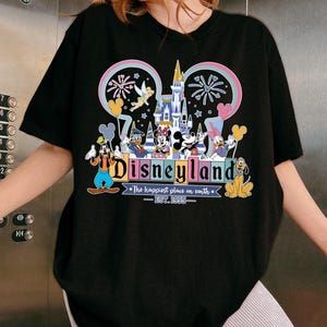 Może przedstawiać: Czarny t-shirt z kolorową grafiką Disneylandu. Projekt zawiera Myszkę Miki, Minnie, Kaczora Donalda, Goofy'ego i Plutona, z napisem "Disneyland" i "The happiest place on earth - EST. 1955."