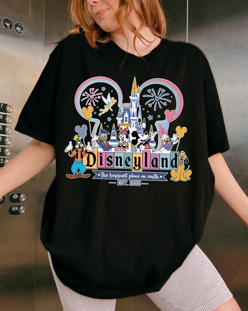 Może przedstawiać: Czarny t-shirt z kolorową grafiką Disneylandu. Projekt zawiera Myszkę Miki, Minnie, Kaczora Donalda, Goofy'ego i Plutona, z napisem "Disneyland" i "The happiest place on earth - EST. 1955."