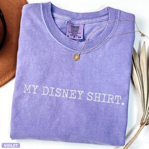May include: A violet Comfort Colors t-shirt with the text "MY DISNEY SHIRT." A gold necklace with a circular pendant rests on the shirt. The shirt is folded, and a brown hat is visible in the background.