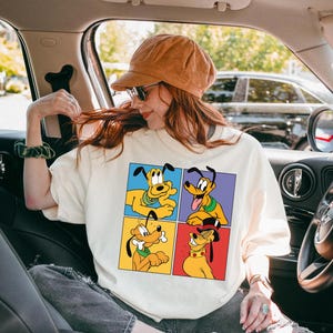 Può includere: T-shirt color crema con una grafica a quattro pannelli di Pluto, il cane dei cartoni animati Disney, in varie pose. La maglietta è indossata da una persona in macchina, con un berretto di velluto a coste marrone e occhiali da sole. Lo sfondo include un'auto e alberi.