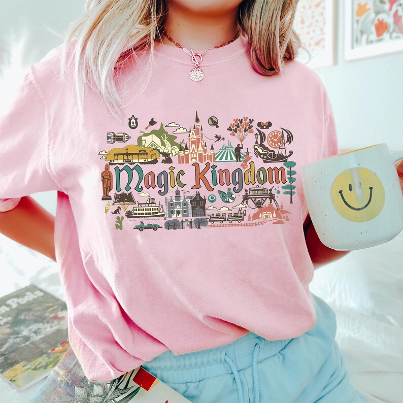 May include: A light pink t-shirt with a colorful graphic of the Magic Kingdom. The graphic includes illustrations of a castle, train, and other park icons. A white mug with a yellow smiley face is held in the hand.