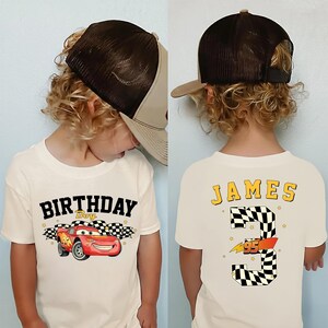 May include: A child wearing a white t-shirt with a graphic of the character Lightning McQueen from the movie Cars, with the text "BIRTHDAY Boy". The back of the shirt has the name "JAMES" and the number "3" with a checkered flag design. The child is also wearing a brown and tan trucker hat.