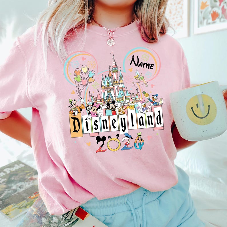 Op de afbeelding: Roze crewneck T-shirt met een Disneyland-afbeelding. De afbeelding toont een kasteel, Disney-figuren en de tekst "Disneyland 2020". Een mok met een smiley wordt in de hand gehouden.