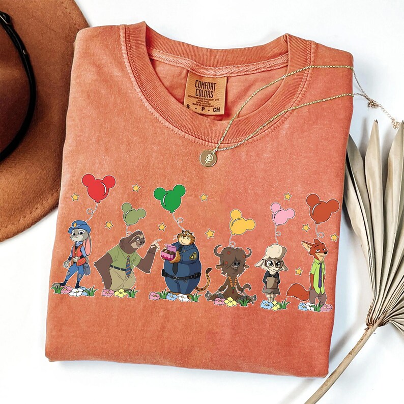 以下が含まれることがあります： オレンジ色のComfort Colors Tシャツ。映画「ズートピア」のキャラクターが風船を持っている漫画のデザイン。 金色のネックレスと茶色の帽子が背景にあります。 Tシャツには小さなComfort Colorsのラベルが付いています。