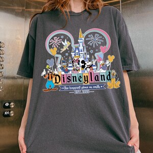 Może przedstawiać: Ciemnoszara koszulka z kolorową grafiką Disneyland. Projekt zawiera logo Disneyland, kultowe postacie, takie jak Myszka Miki, oraz napis "The happiest place on earth - EST. 1955."