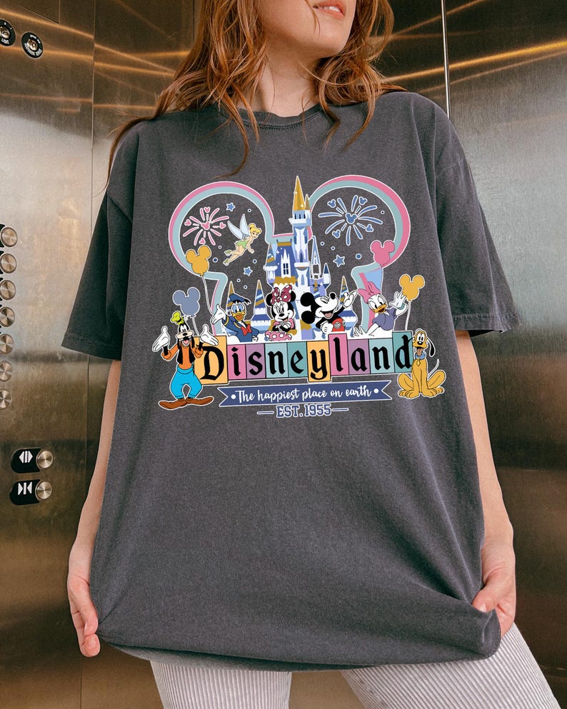 Może przedstawiać: Ciemnoszara koszulka z kolorową grafiką Disneyland. Projekt zawiera logo Disneyland, kultowe postacie, takie jak Myszka Miki, oraz napis "The happiest place on earth - EST. 1955."