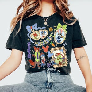 May include: Black t-shirt featuring colorful illustrations of characters from the animated film Zootopia. The design includes framed portraits, a carrot, and the word "Zootopia" in a decorative font. The shirt has a vintage, worn look.