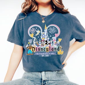 Może przedstawiać: Niebieska koszulka z grafiką Disneyland. Grafika zawiera zamek, Myszka Miki i Myszka Minnie, Kaczor Donald, Kaczka Daisy, Goofy i Pluto. Tekst brzmi "Disneyland" i "The happiest place on earth - EST. 1955."