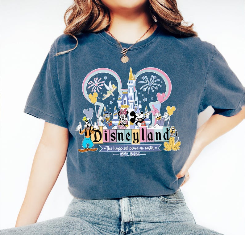 Może przedstawiać: Niebieska koszulka z grafiką Disneyland. Grafika zawiera zamek, Myszka Miki i Myszka Minnie, Kaczor Donald, Kaczka Daisy, Goofy i Pluto. Tekst brzmi "Disneyland" i "The happiest place on earth - EST. 1955."