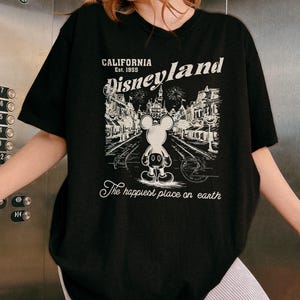 May include: Black t-shirt featuring a graphic of Mickey Mouse in front of Disneyland. The design includes the text "CALIFORNIA Est. 1955 Disneyland" and "The happiest place on earth" in white. The shirt has a crew neck and short sleeves.