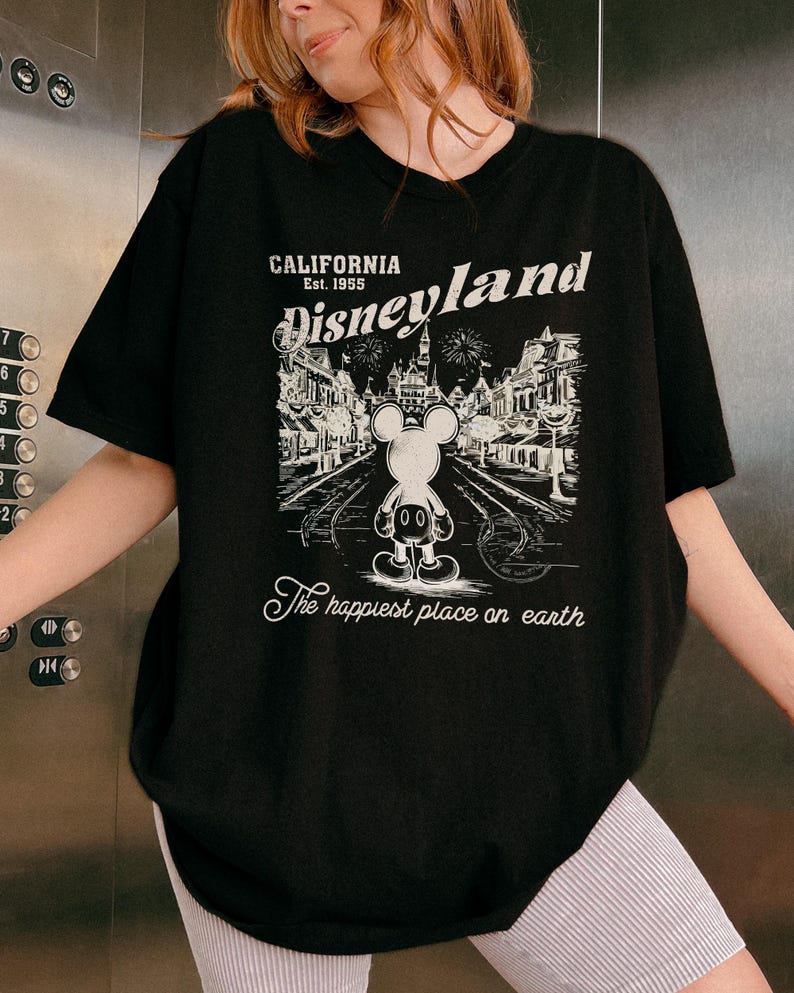 May include: Black t-shirt featuring a graphic of Mickey Mouse in front of Disneyland. The design includes the text "CALIFORNIA Est. 1955 Disneyland" and "The happiest place on earth" in white. The shirt has a crew neck and short sleeves.