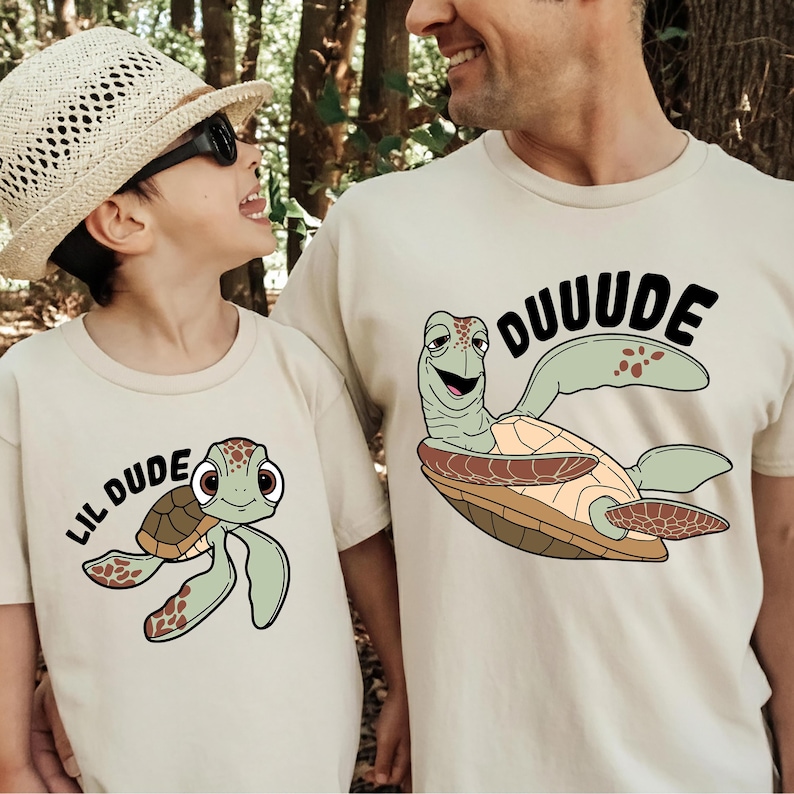 May include: Beige t-shirts with turtle graphics. One shirt says "DUUUUDE" and the other says "LIL DUDE". The turtle illustrations are green, brown, and tan, with black outlines. The shirts are worn by a child and an adult.