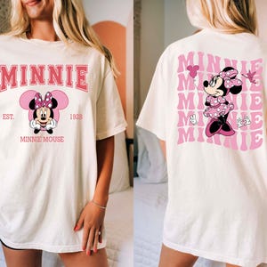 May include: Cream-colored t-shirt featuring a Minnie Mouse graphic. The front design includes the word "MINNIE" in pink, along with an illustration of Minnie Mouse and the text "EST. 1928 MINNIE MOUSE." The back of the shirt has a repeating pink "MINNIE" design and a Minnie Mouse illustration.