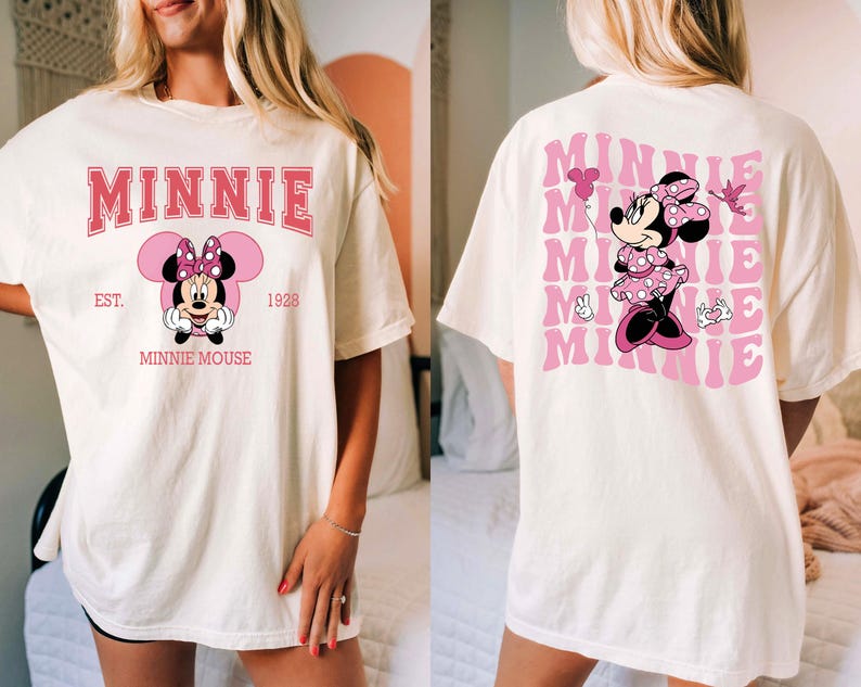 May include: Cream-colored t-shirt featuring a Minnie Mouse graphic. The front design includes the word "MINNIE" in pink, along with an illustration of Minnie Mouse and the text "EST. 1928 MINNIE MOUSE." The back of the shirt has a repeating pink "MINNIE" design and a Minnie Mouse illustration.