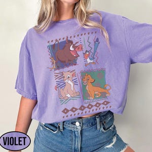 May include: Violet-colored t-shirt featuring a graphic of characters from The Lion King. The design includes images of Timon, Pumbaa, Simba, and Nala, set against a geometric pattern. The word "VIOLET" is printed on the bottom left.