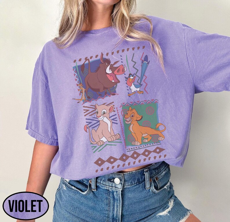 May include: Violet-colored t-shirt featuring a graphic of characters from The Lion King. The design includes images of Timon, Pumbaa, Simba, and Nala, set against a geometric pattern. The word "VIOLET" is printed on the bottom left.