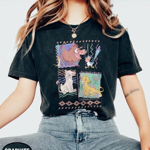 May include: Graphite-colored t-shirt featuring a colorful graphic of characters from The Lion King. The design includes images of Timon, Pumbaa, Simba, and Zazu. The shirt has a classic crew neck and short sleeves.
