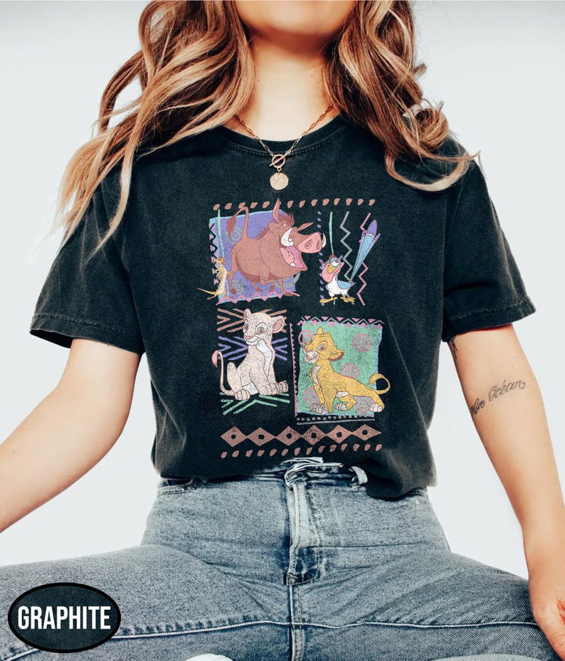 May include: Graphite-colored t-shirt featuring a colorful graphic of characters from The Lion King. The design includes images of Timon, Pumbaa, Simba, and Zazu. The shirt has a classic crew neck and short sleeves.
