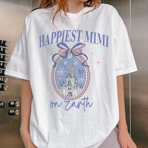 K&ouml;nnte beinhalten: Wei&szlig;es T-Shirt mit dem Text "Happiest Mimi on Earth" in hellvioletter Schrift. Das Shirt zeigt eine Grafik eines Schlosses in einem dekorativen Oval mit einer Schleife und Feuerwerk.