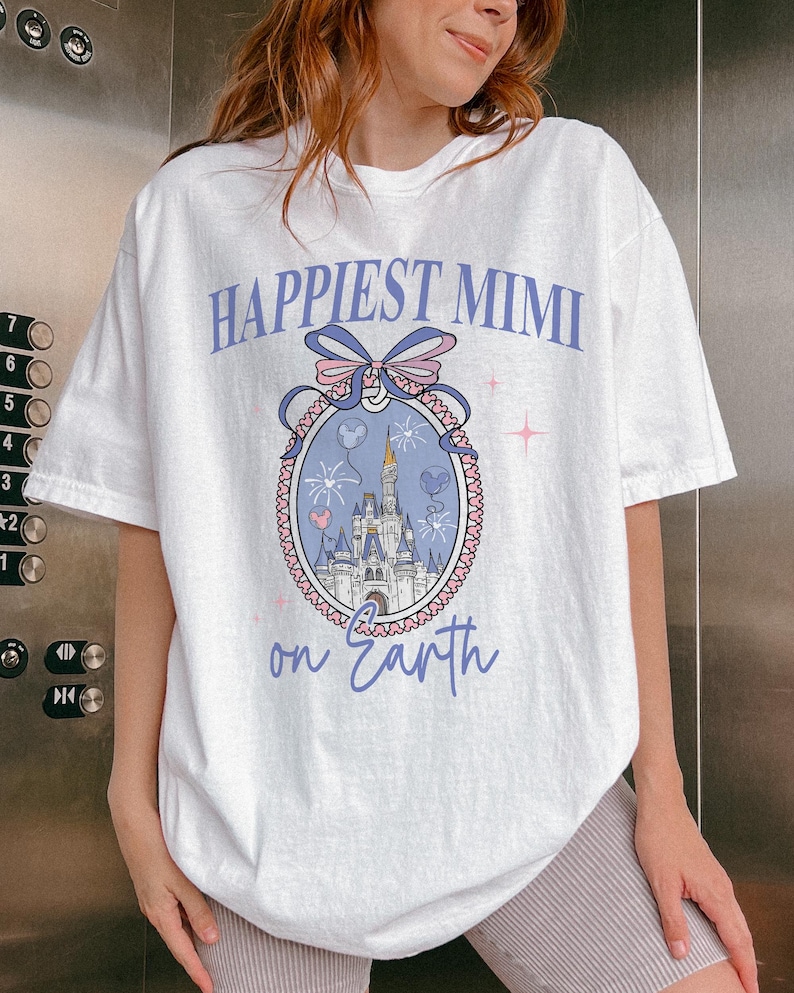 K&ouml;nnte beinhalten: Wei&szlig;es T-Shirt mit dem Text "Happiest Mimi on Earth" in hellvioletter Schrift. Das Shirt zeigt eine Grafik eines Schlosses in einem dekorativen Oval mit einer Schleife und Feuerwerk.
